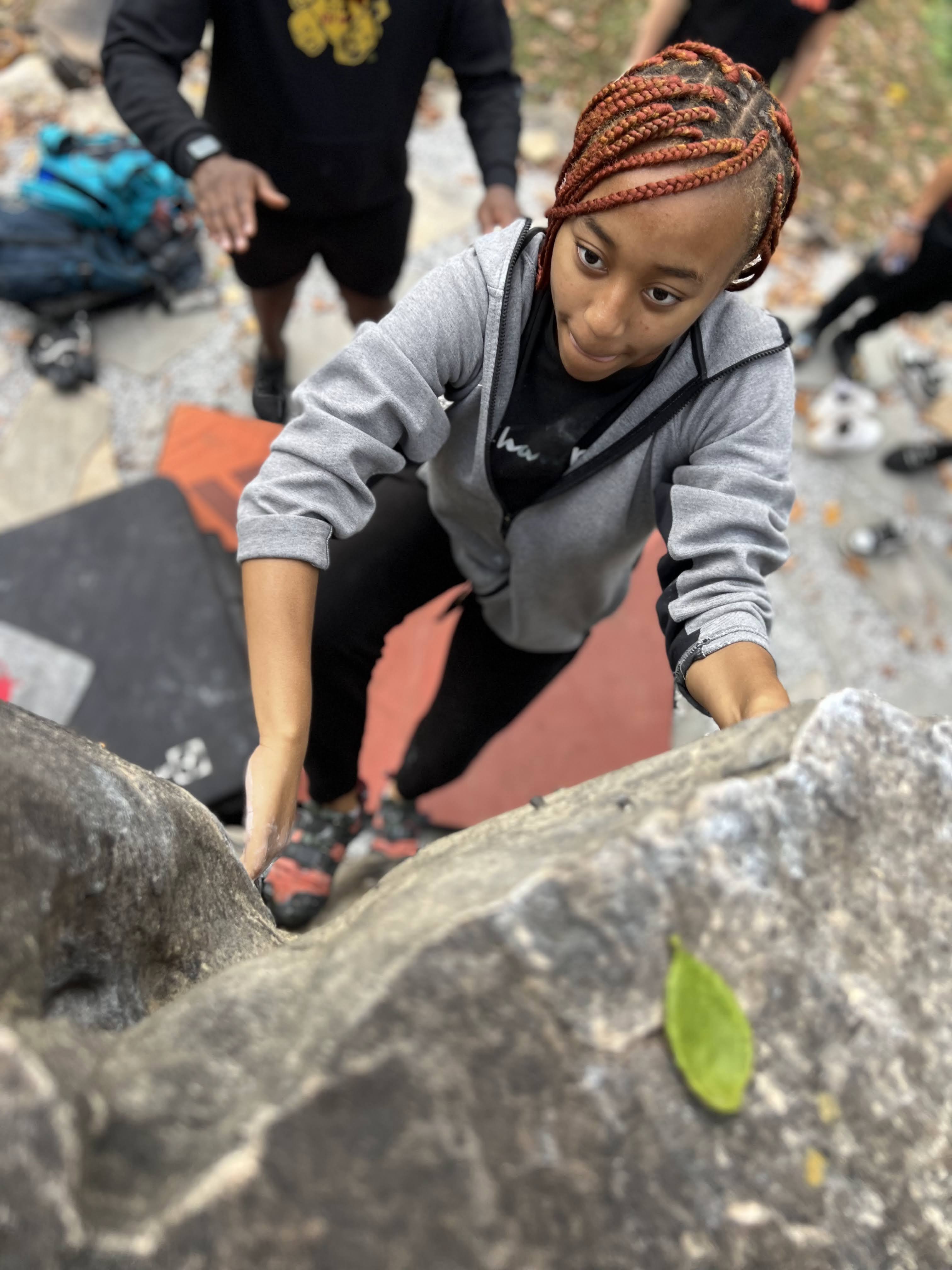 A student rock climbs with arms below her offering support